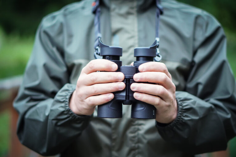 Close up of a man dressed in a hunter jacket holds binoculars in his hand.